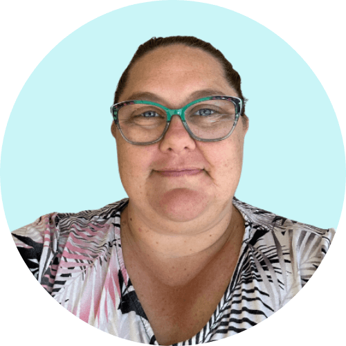 Image of a woman wearing a floral top and green framed glasses. Her dark hair is tied back, and she is smiling with her mouth closed.