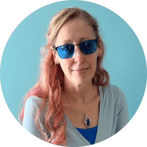 Image of a woman with long blonde and pink hair, wearing bright blue glasses and smiling with her mouth closed.