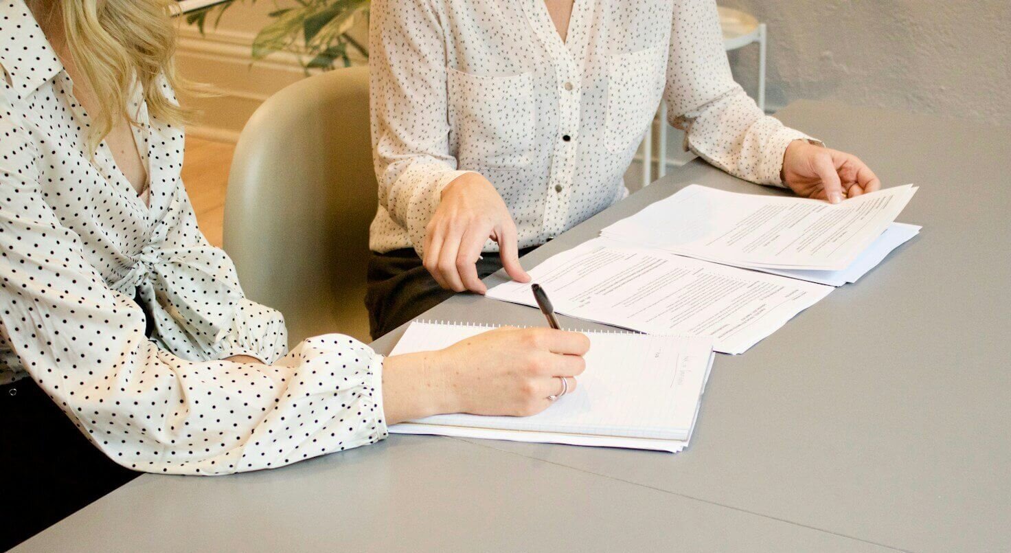 woman signing on white printer paper beside woman about to touch the documents